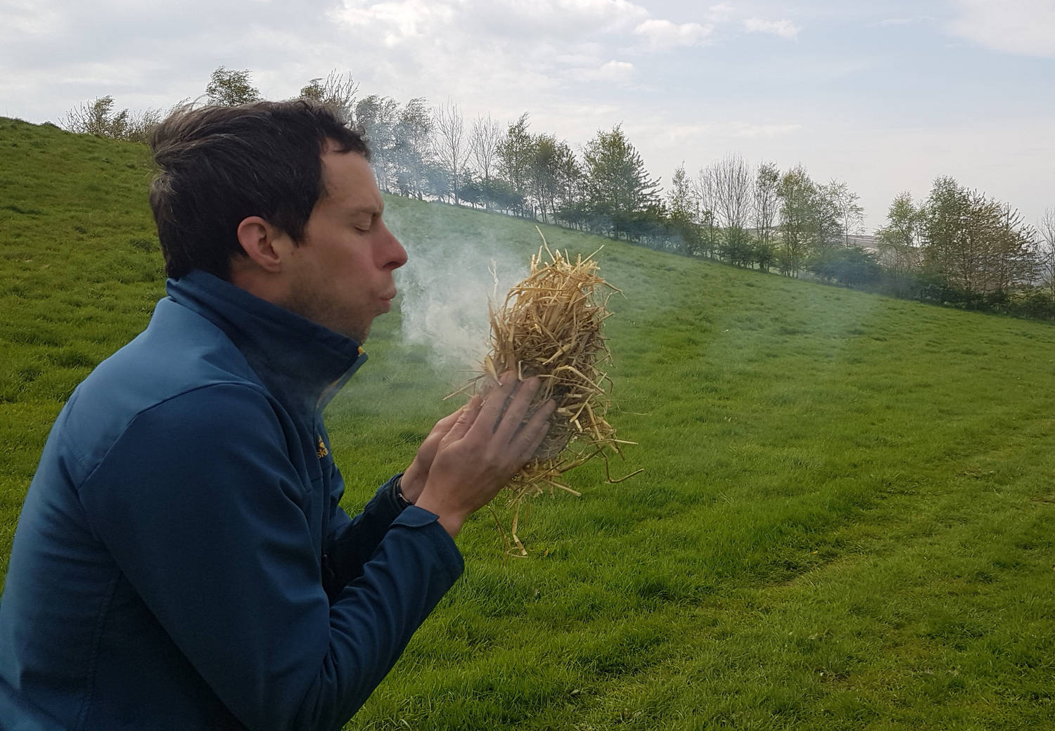 A photograph of a man blowing ona tinder bundle in a field.