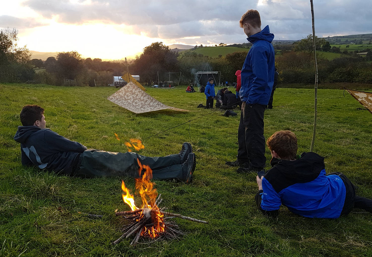 Three people relaxing outside a trap shelter in front of a  camp fire in a field